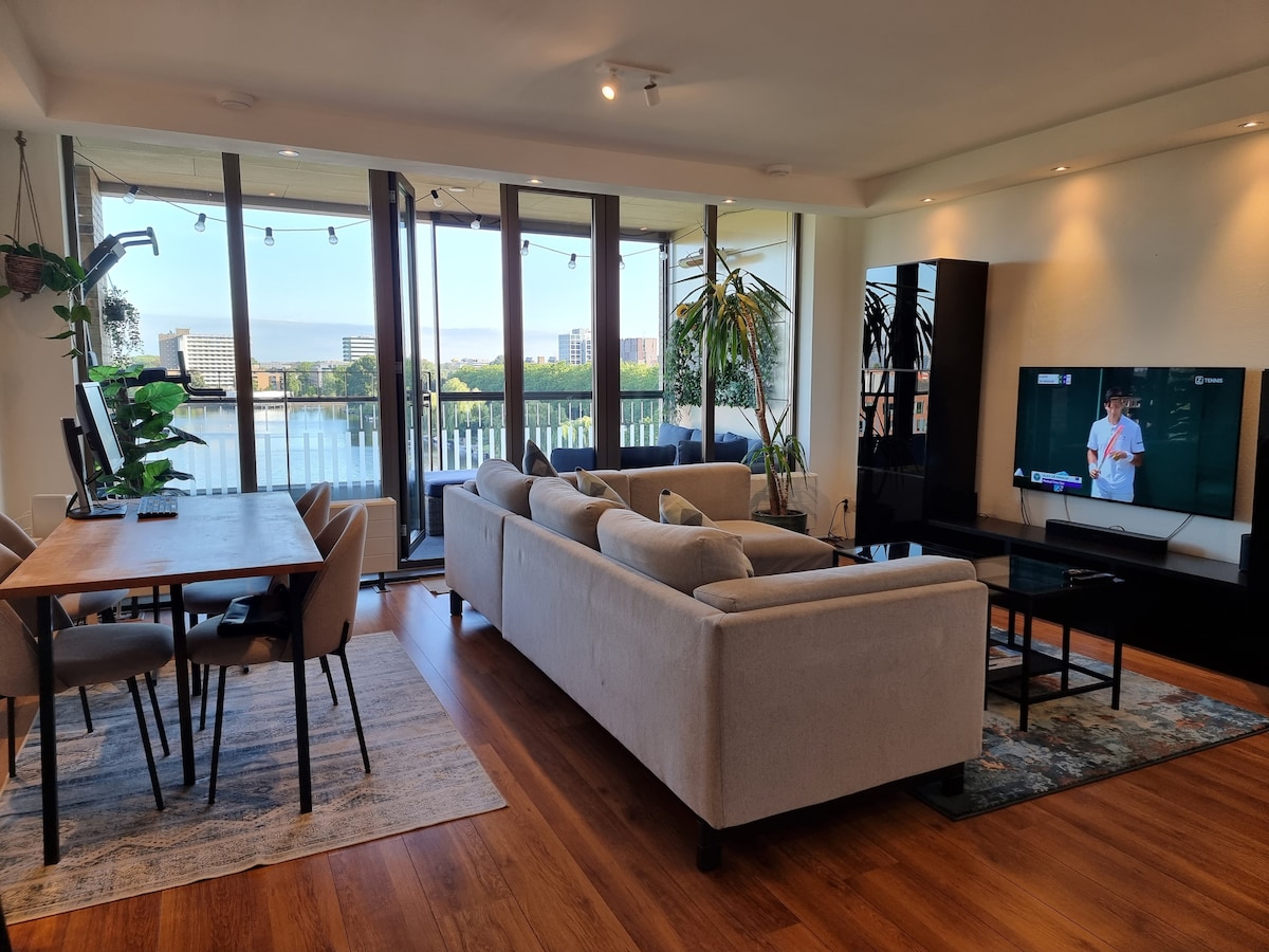 The living area features a light-colored sectional sofa facing a large flatscreen TV. A dining table with several chairs is positioned nearby, with ample natural light streaming in through expansive glass doors that open to a balcony overlooking the water.