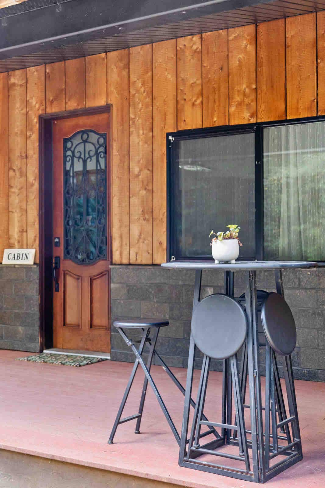 An entrance is framed by warm wooden siding and a decorative door with an intricate design. A tall table with four round stools stands nearby, accompanied by a small potted plant. Large windows allow natural light to fill the space.
