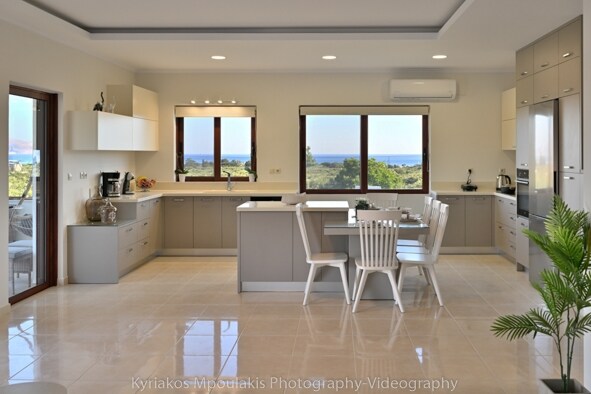 A modern kitchen features light-colored cabinetry and a central island with seating. Two large windows allow natural light to illuminate the space and offer views of distant mountains and the sea. The tiled floor reflects the bright ambiance, enhancing the kitchen's spaciousness.