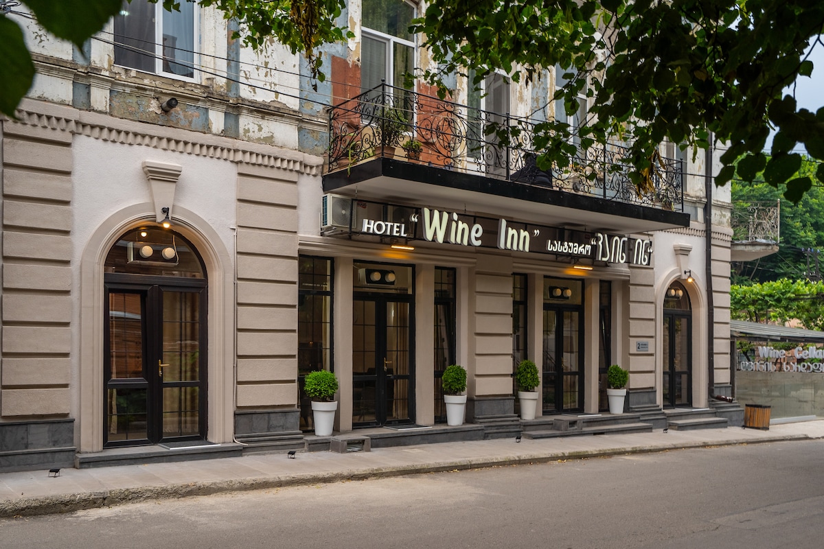 The hotel exterior showcases a blend of classic architectural elements, with decorative moldings and large windows framed by elegant black wrought iron. Symmetrical planters with greenery add a touch of nature to the entrance, while the signage identifies the establishment.