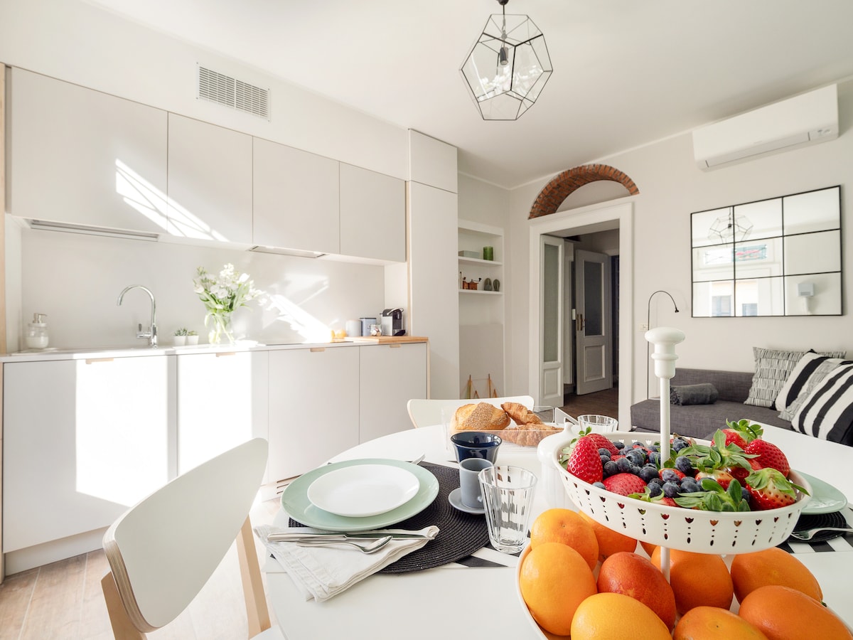 A well-lit kitchen and dining area features a round table adorned with a bowl of fresh fruits, including oranges, strawberries, and blueberries. Modern cabinetry and appliances are visible in the background, complemented by decorative touches. A comfortable living space is seen through an open doorway.