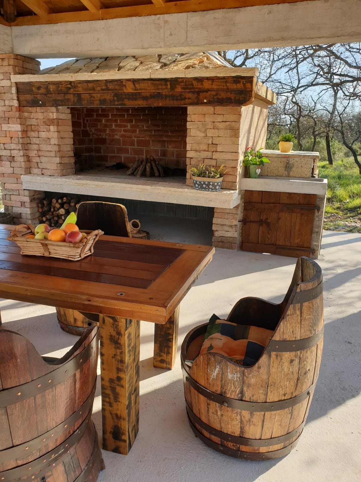 An outdoor BBQ area is presented with a stone fireplace made of brick, accompanied by a wooden dining table. Unique barrel-style chairs are arranged around the table, with a basket of fresh fruits placed prominently at the center. A small garden area is visible in the background.