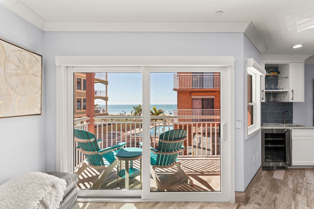A bright and airy living area opens to a balcony with two turquoise chairs facing a view of the ocean. Sunlight streams through the sliding glass door, enhancing the coastal atmosphere. The nearby kitchen features modern cabinetry and a beverage refrigerator.
