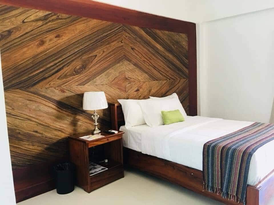 A cozy sleeping area is highlighted by a rich wooden accent wall featuring a chevron pattern. A spacious bed is made with crisp white linens and a small green accent pillow. A wooden bedside table and lamp provide functionality, complementing the overall design.