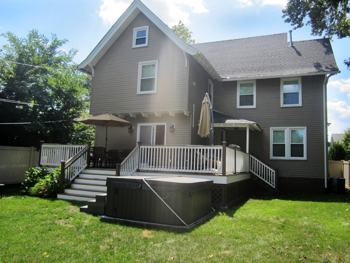 A spacious backyard is showcased, featuring a roomy deck with an outdoor dining area and an umbrella. A hot tub is positioned nearby, and well-maintained grass surrounds the area. The house façade is visible, highlighting multiple windows and a second-level balcony.