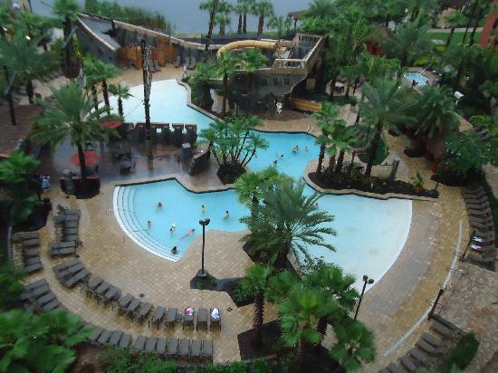 An expansive pool area is surrounded by lush palm trees, featuring multiple swimming pools interconnected by winding pathways. Lounge chairs are arranged around the pools, and guests can be seen enjoying the water. A waterfall structure adds to the tropical ambiance.