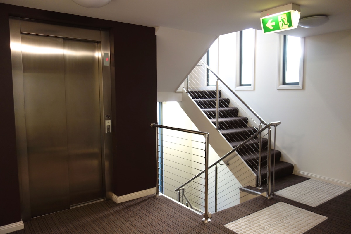 An area features an elevator positioned next to a set of stairs, with a modern railing providing safety. Natural light enters through large windows, enhancing the spaciousness of the hallway. Clear signage indicates the direction of the emergency exit.