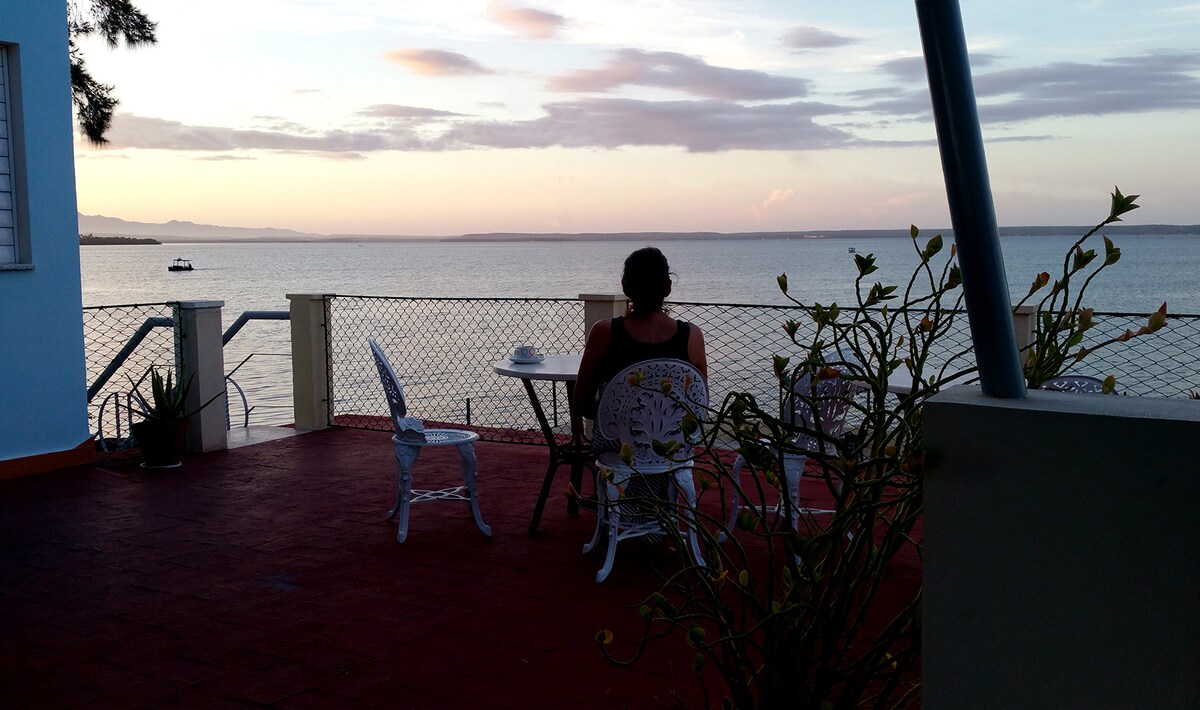 A person is seated at a small table on a terrace overlooking the water, with boats visible in the distance. The scene is framed by a protective railing and surrounded by subtle greenery, creating a peaceful setting during sunset.