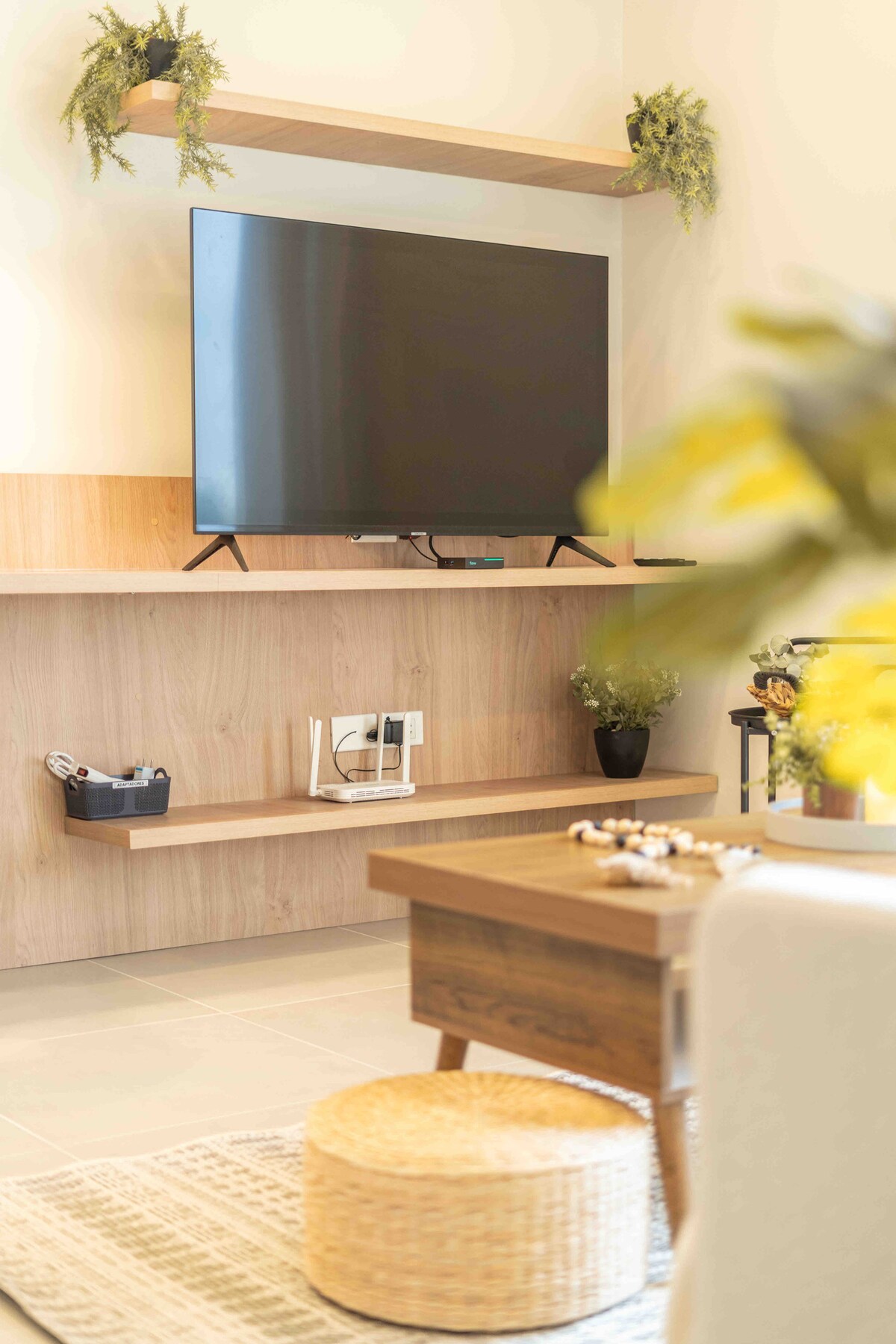 A TV mounted on a light wooden shelf is positioned above a wooden console. A small table and a decorative stool are present below, complemented by potted plants on the shelf. Soft lighting enhances a cozy yet functional living area.
