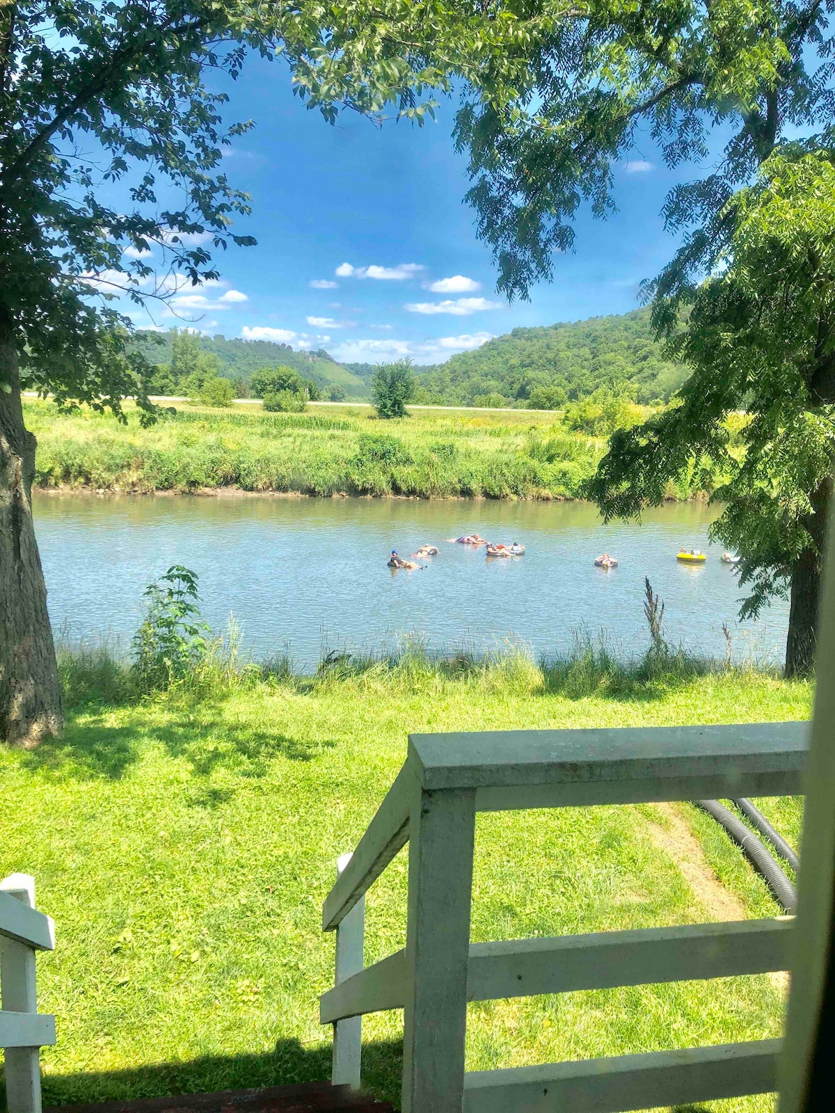 A serene view of the Upper Iowa River is captured, showcasing gentle ripples and colorful kayaks floating along the water. Lush greenery surrounds the riverbank, with trees providing shade and a staircase leading down to the river, enhancing the natural setting.