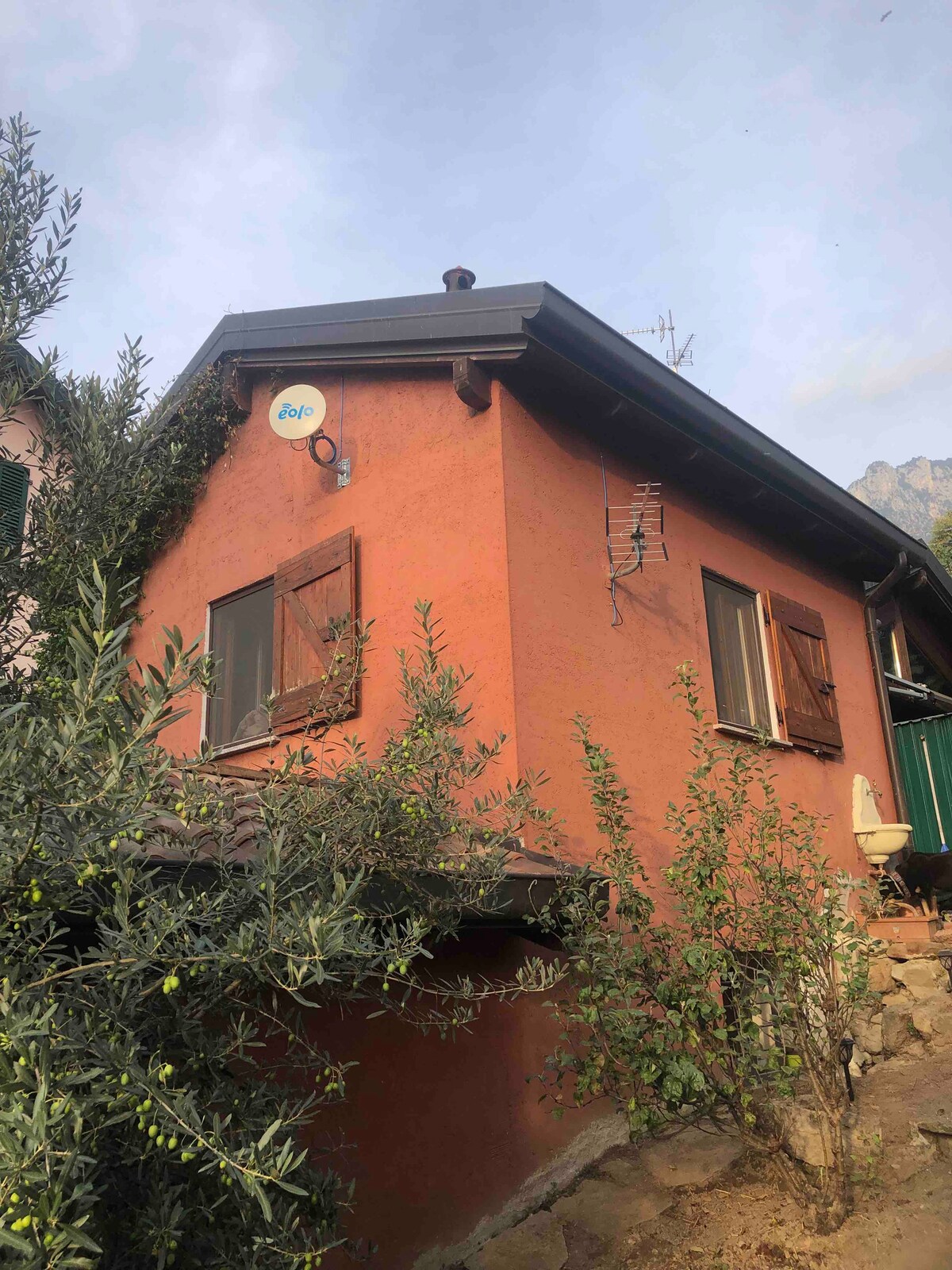 The exterior of a two-story orange house is visible, featuring wooden shutters and a satellite dish. Olive trees are seen in the foreground, and surrounding mountains can be glimpsed in the background under a cloudy sky.