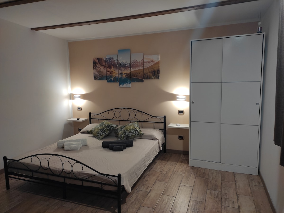 A spacious sleeping area features a metal bed frame with a light-colored bedding set. A series of mountain-themed artworks adorns the wall above the bed. Nightstands with lamps are positioned on either side, and a white wardrobe is available for storage.