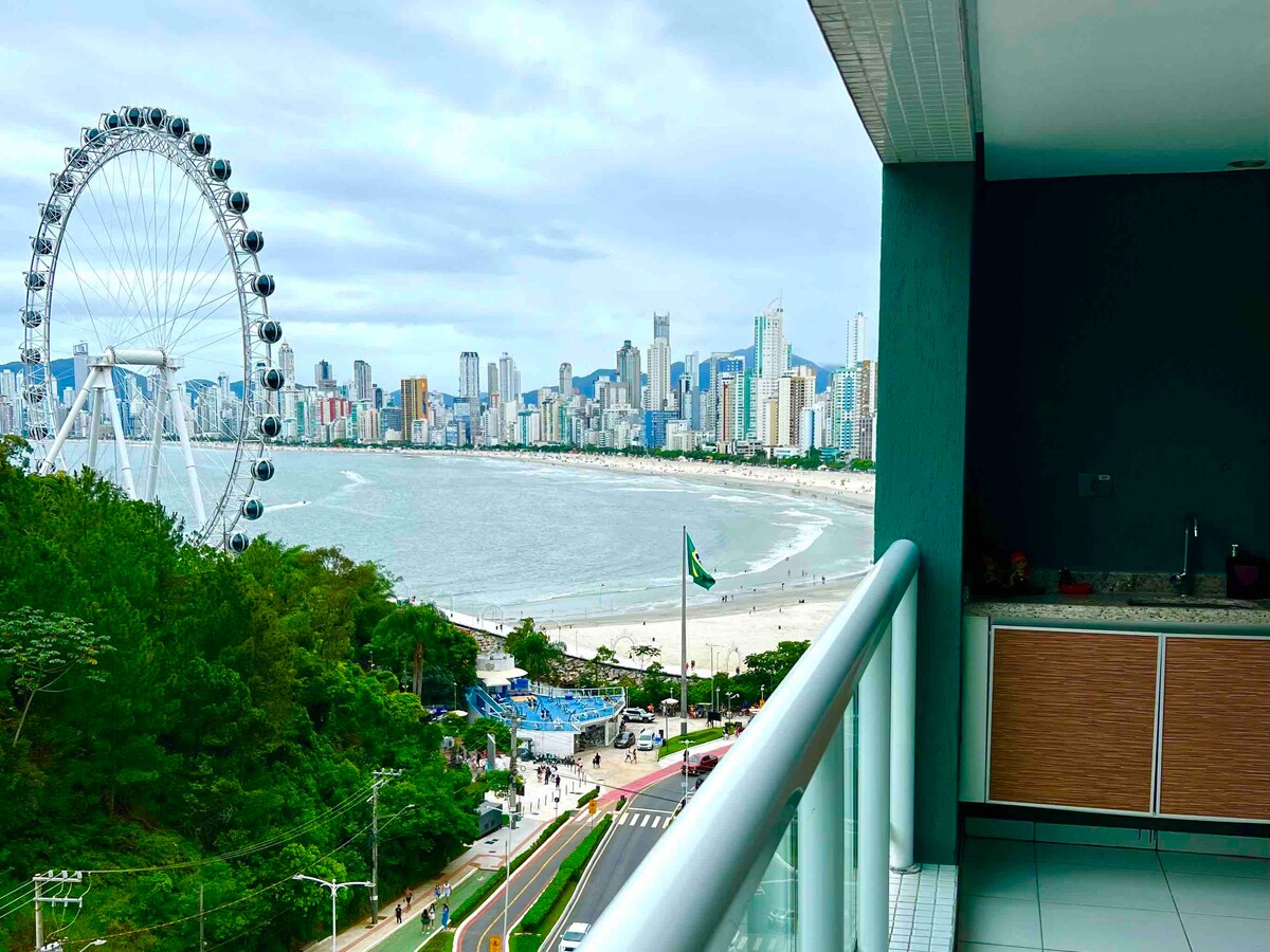 A spacious balcony offers stunning views of the coastline and the iconic Ferris wheel of Balneário Camboriú. The vibrant city skyline is visible in the background, with lush greenery framing the scene. The area below features sandy beaches and walking paths along the shore.
