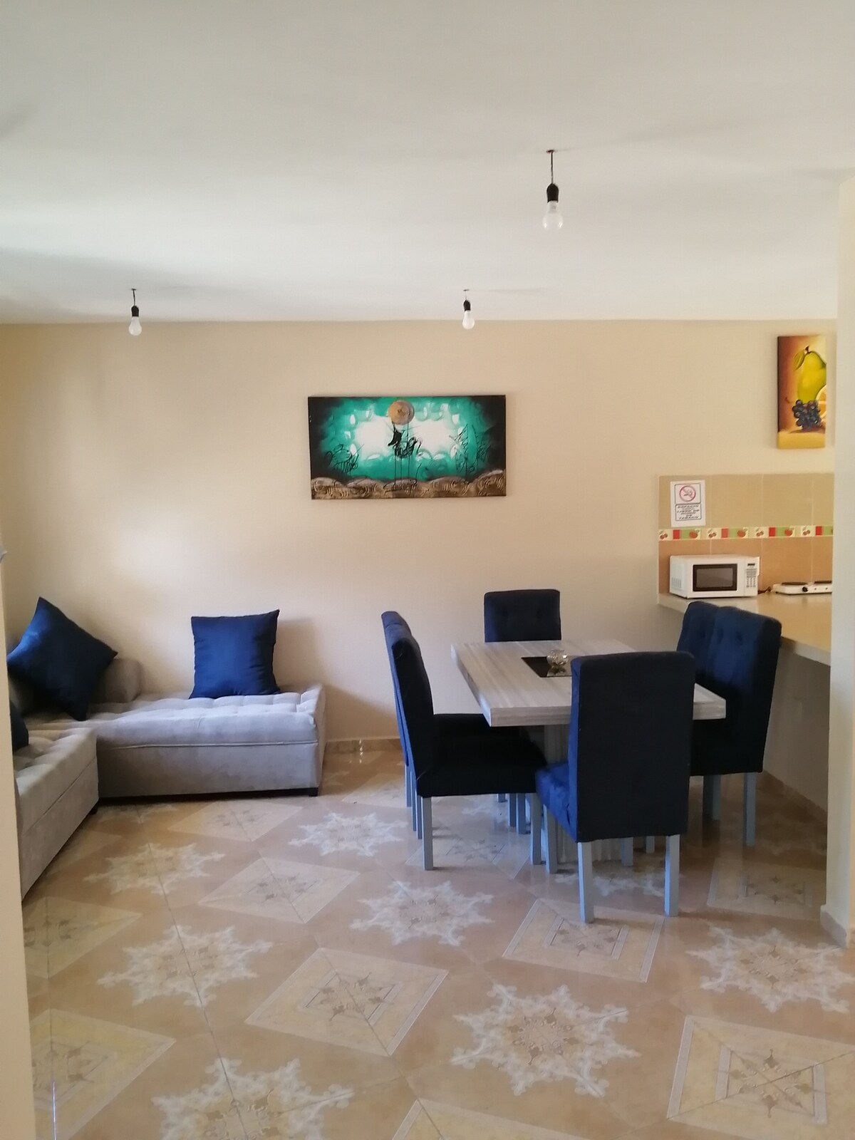 A dining area features a table surrounded by dark chairs, complemented by a soft gray couch adorned with blue pillows. Light-colored walls enhance the space, and a vibrant piece of wall art is displayed above the dining area, contributing to the overall ambiance.