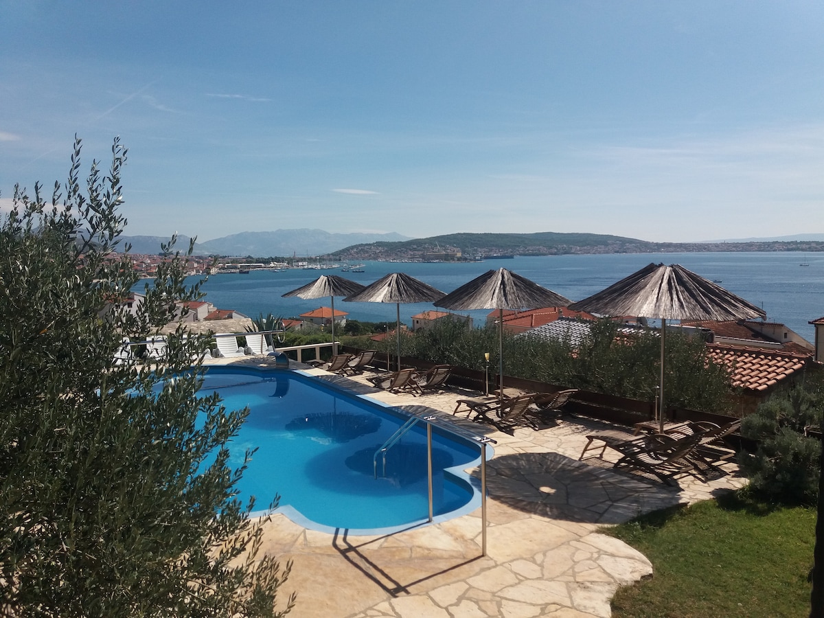 A crystal-clear swimming pool is surrounded by loungers and umbrellas, offering views of the sea and distant hills. Olive trees provide greenery in the foreground, enhancing the serene setting under a clear blue sky.