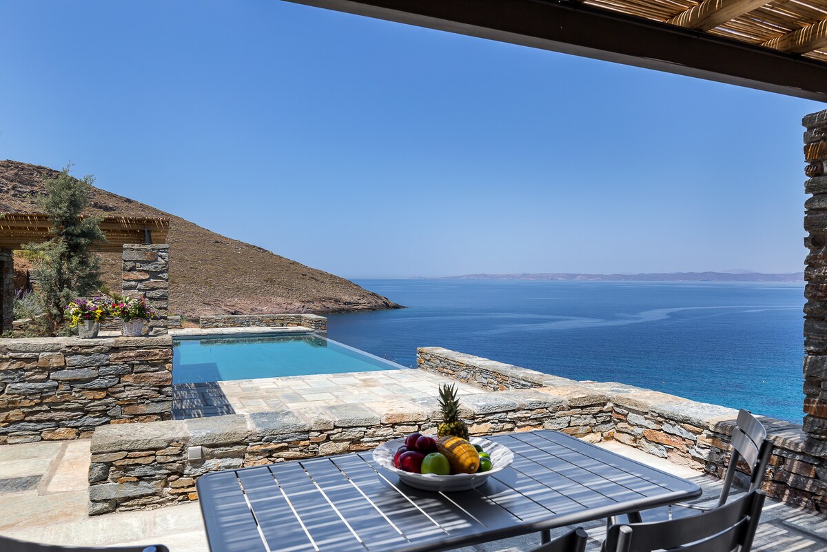 A shaded outdoor dining area features a black table set with a colorful assortment of fruits. Beyond, a serene infinity pool appears to blend with the blue sea, while a rocky hillside frames the view under a clear sky.