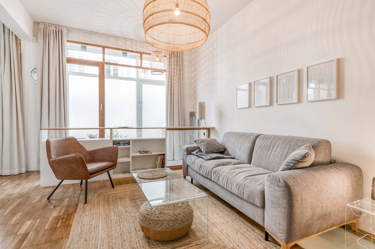 A bright living area features a comfortable grey sofa paired with a stylish armchair. A glass coffee table sits on a woven rug, while large windows allow natural light to fill the space. Simple artwork adorns the walls, enhancing the clean and airy atmosphere.