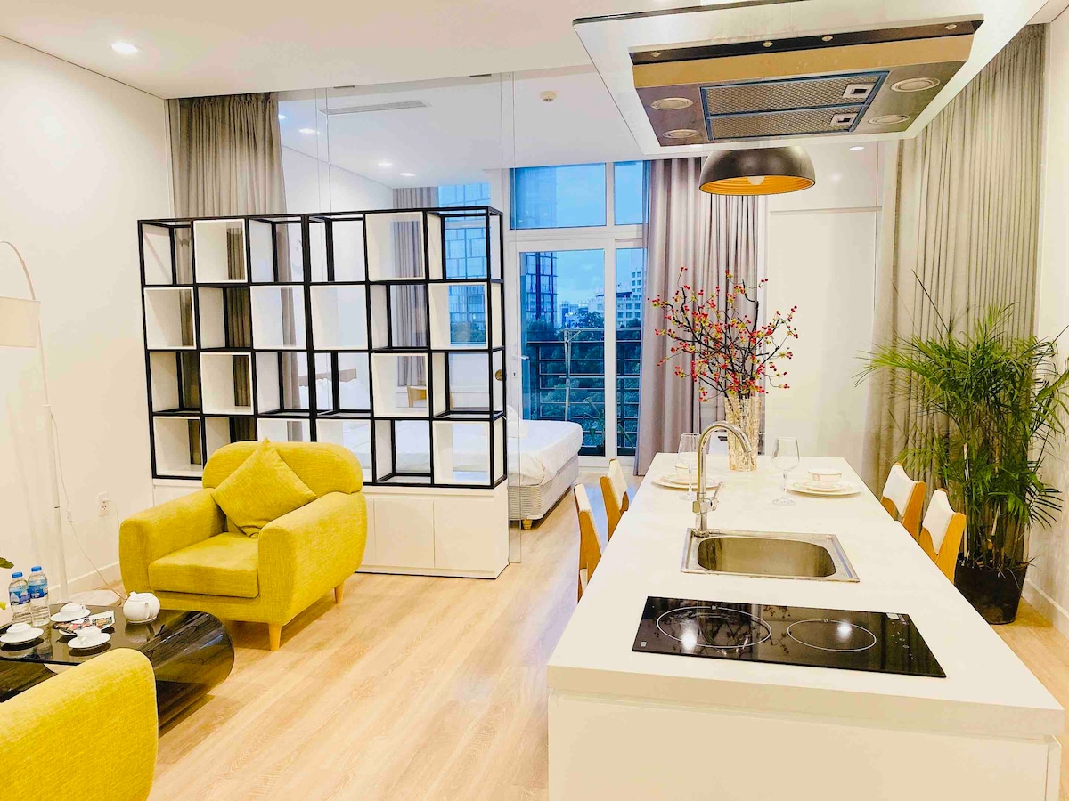 A modern open-concept space features a sleek kitchen with an island, complete with a stovetop and sink. A dining area is set with tableware. Yellow armchairs provide seating, while a room divider reveals a sleeping area in the background, and large windows allow natural light into the room.