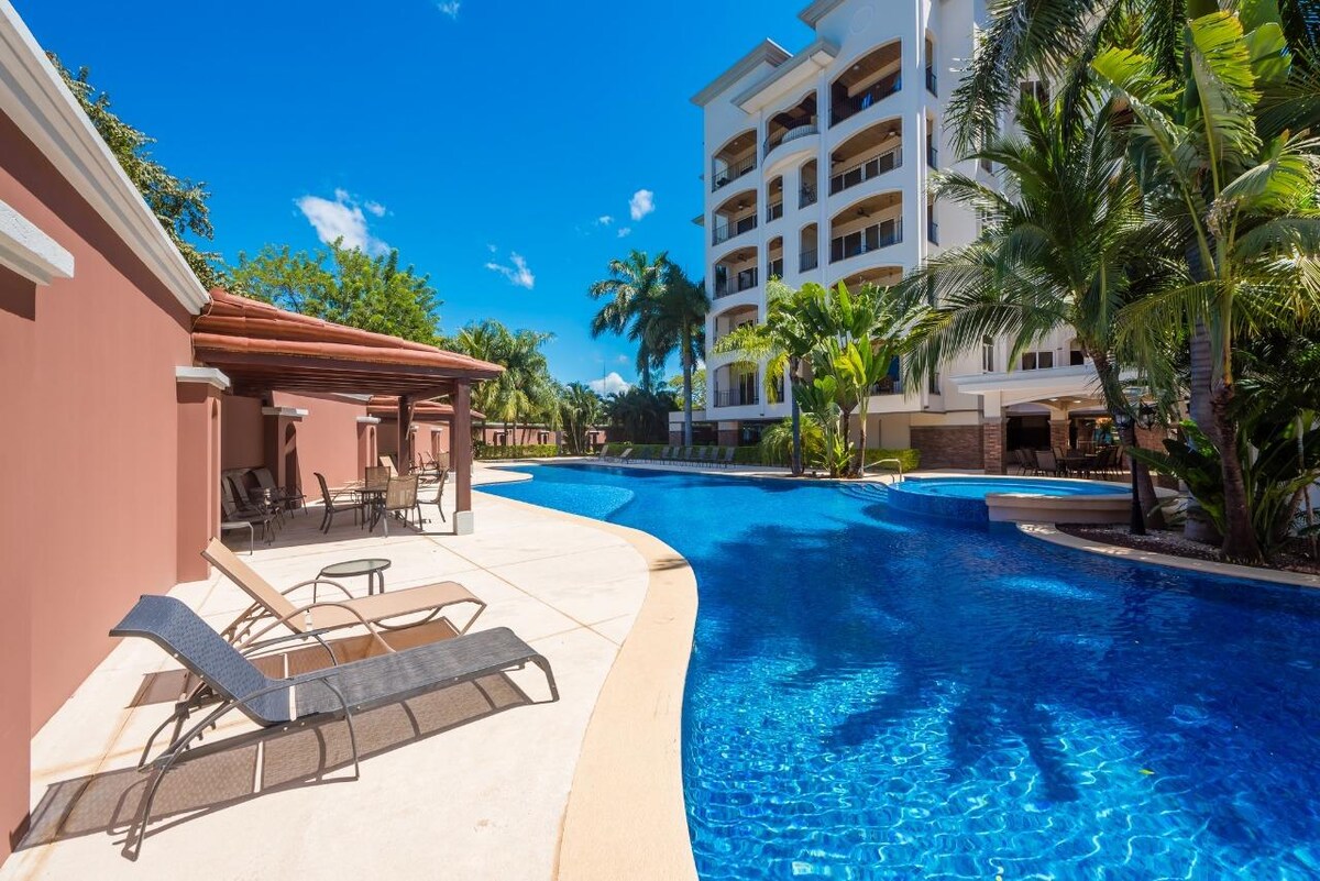 A spacious pool area is surrounded by tropical landscaping and palm trees. Lounge chairs are positioned along the water's edge, inviting relaxation. The pool features a curved design with various depths and is bathed in natural sunlight, creating a bright and refreshing atmosphere.