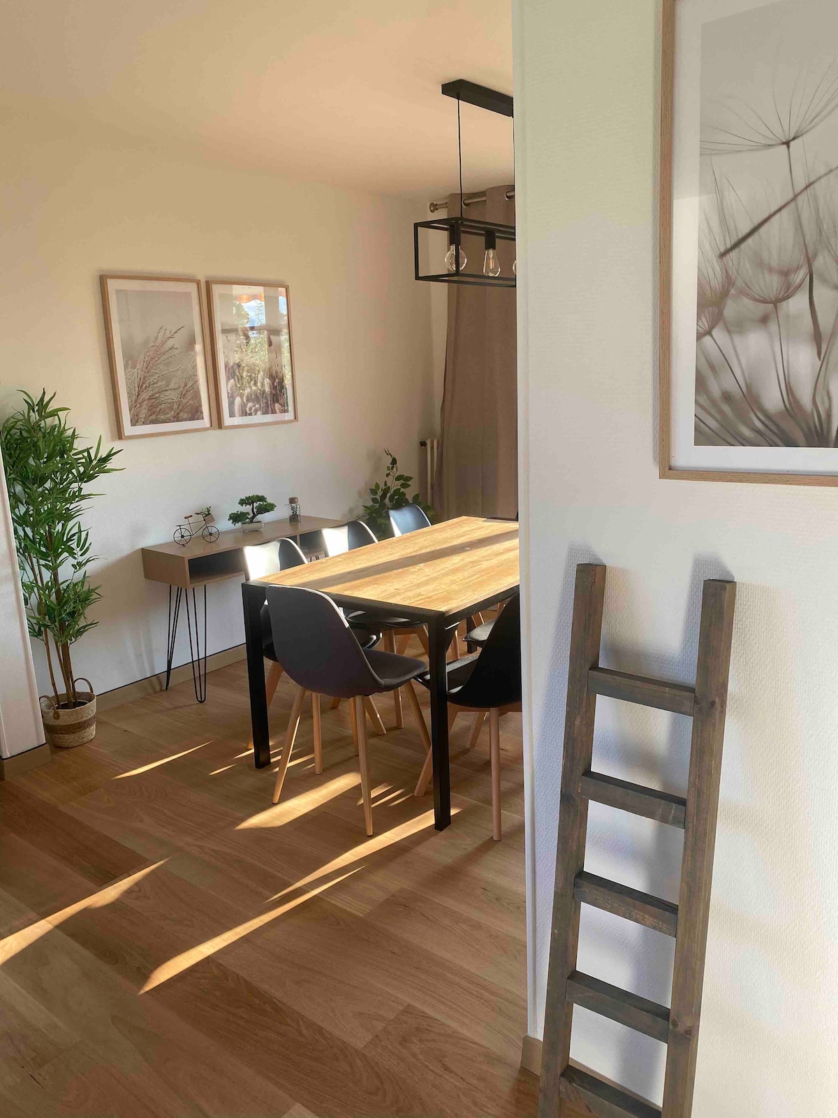 A dining area is visible, featuring a wooden table surrounded by six chairs. Natural light enters through a nearby window, highlighting the warm wood floor. Plants are placed around the space, and framed artwork adorns the walls, contributing to the overall serene ambiance.