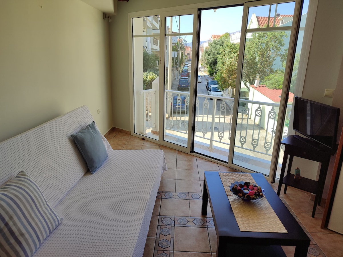 The living area features a light-colored sofa with a decorative pillow, situated beside large glass doors that open to a balcony. A small coffee table is positioned in front, and a television stands on a console nearby. Natural light fills the space.