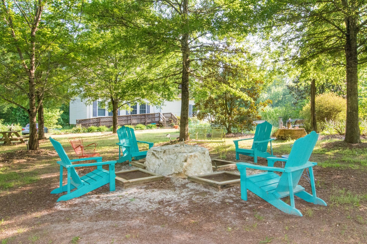 A fire pit is surrounded by four turquoise Adirondack chairs and features a stone seating area. Lush greenery from surrounding trees provides shade, enhancing the outdoor space's inviting atmosphere. In the background, a cabin is visible, blending into the natural setting.