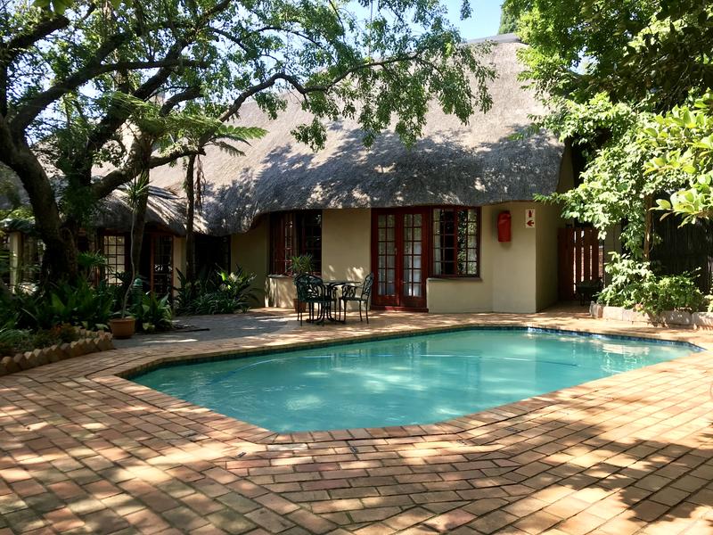 A refreshing swimming pool is surrounded by a spacious patio made of interlocking bricks. Lush greenery frames the area, with a thatched-roof chalet visible in the background. A dining set is located nearby, providing an inviting space for outdoor meals.