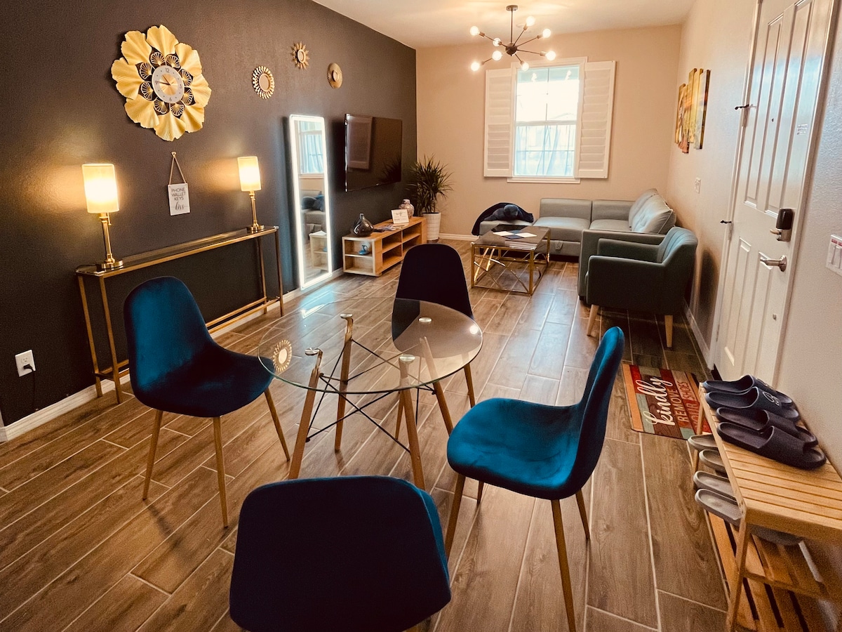 The living area features a modern design with a glass table surrounded by four upholstered chairs. A comfortable sofa is positioned nearby, facing a wall-mounted TV. Decorative elements, such as a flower design and ambient lighting, enhance the space, while natural light filters in through the window.