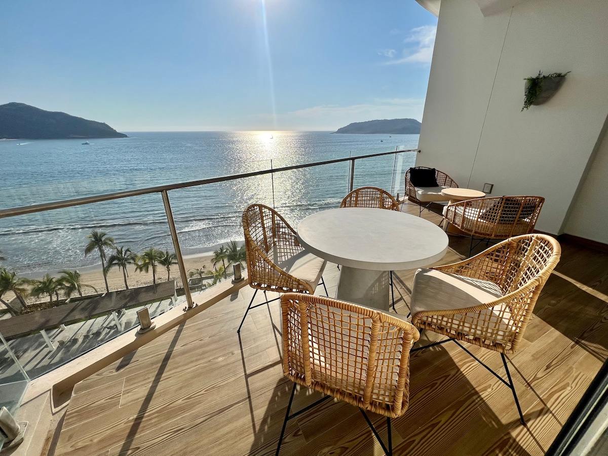 A balcony overlooks the beach, featuring a round table surrounded by four wicker chairs. The ocean sparkles under sunlight, with distant mountains framing the view. Lush palm trees and sandy beach are visible below, creating a serene coastal setting.