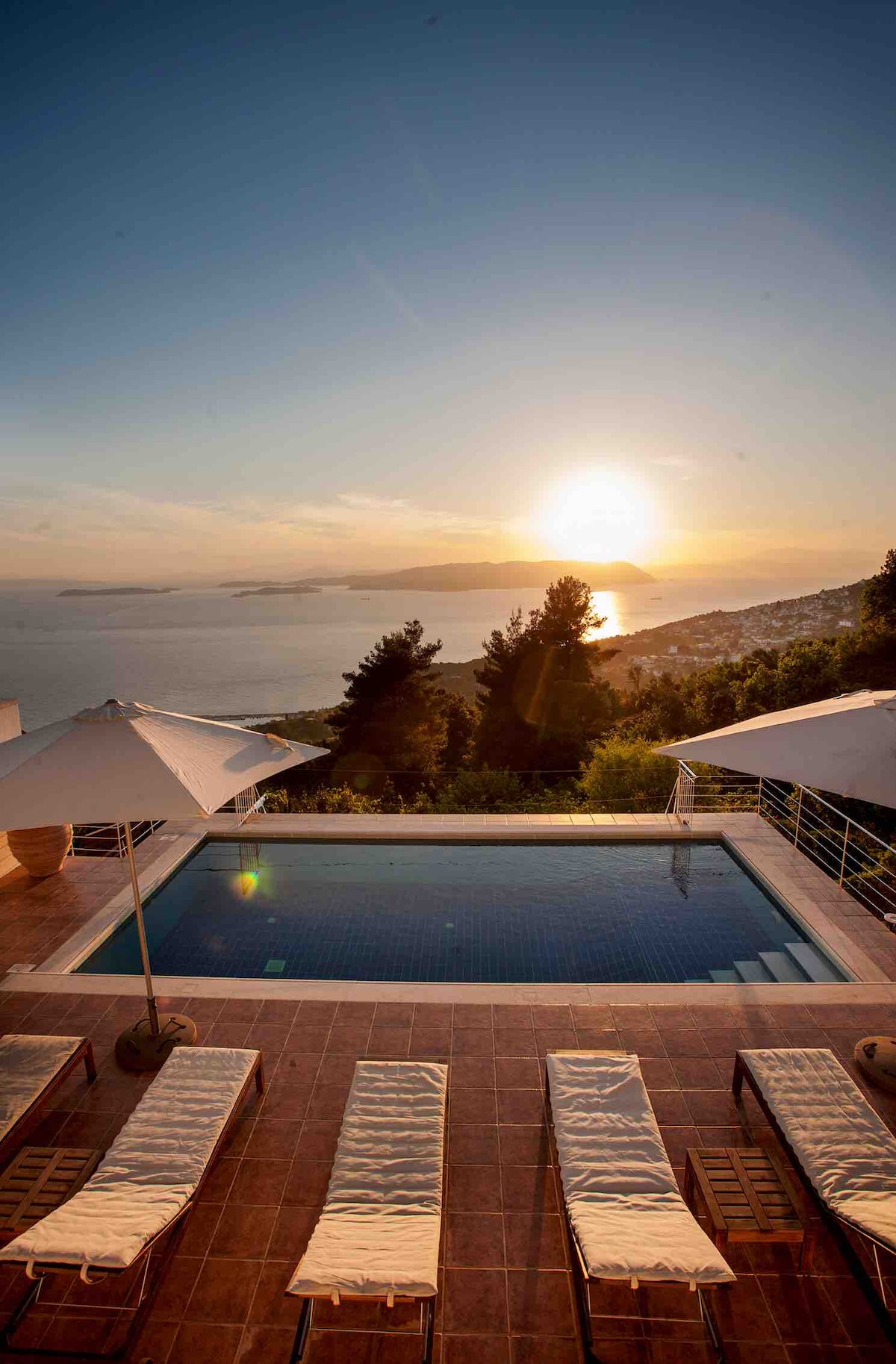 A serene swimming pool area is showcased with sun loungers lined along the edge. Large umbrellas provide shade, while the horizon displays a beautiful sunset over the sea. Hills and distant islands are visible, contributing to the peaceful ambiance.