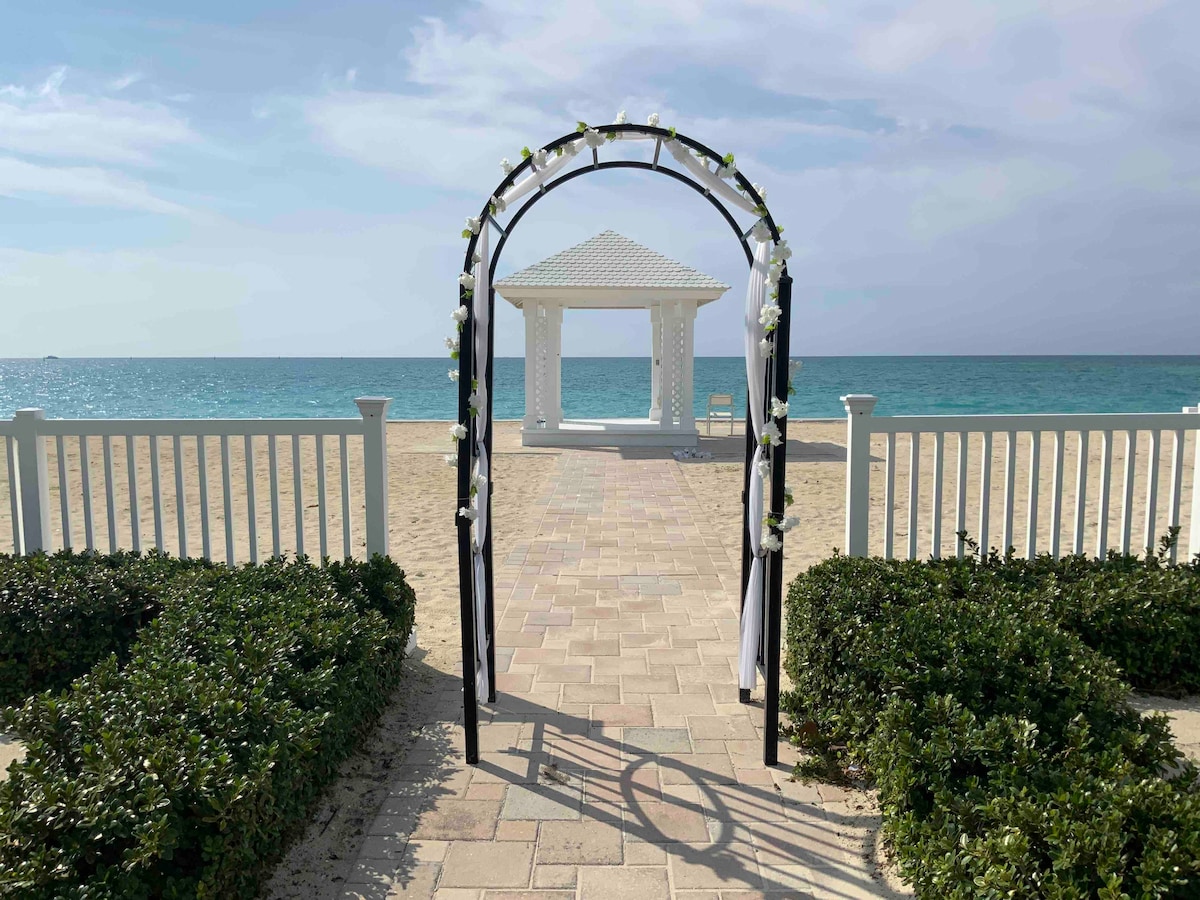 An arch adorned with white flowers frames a pathway leading to a covered pavilion beside the beach. The sandy area extends towards the turquoise water, with a low white fence and green shrubs on either side enhancing the coastal setting.