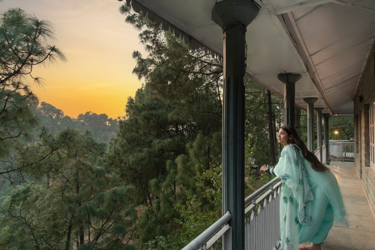 A spacious balcony is viewed, framed by tall pillars and surrounded by lush green pine trees. Soft sunlight casts a warm glow over the landscape as the sky transitions from day to night, creating a serene atmosphere.