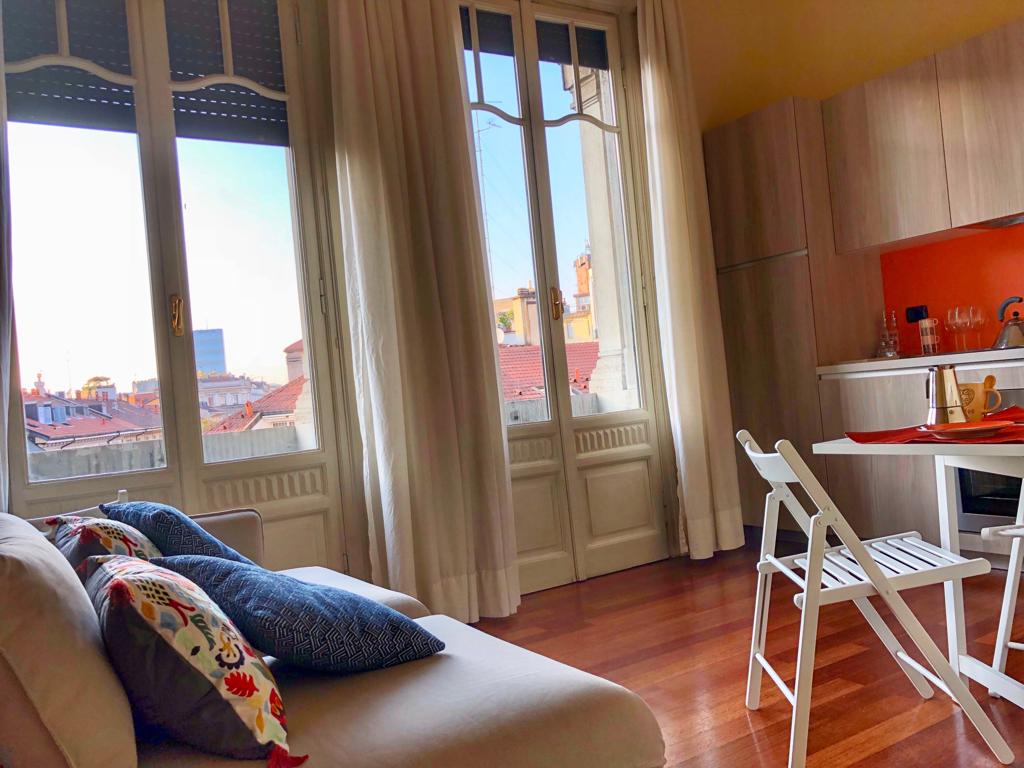 A bright living area is visible, featuring a soft sofa with colorful cushions. Light streams in through large windows, highlighting polished wooden floors. A small dining table with chairs and an elegant kitchen setup can be seen in the background, complemented by warm-toned walls.