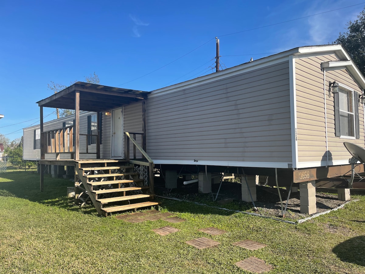 A well-maintained trailer is depicted with a wooden porch featuring steps leading up to the entrance. The exterior is light-colored, complemented by a clear blue sky. Surrounding grassy areas are visible, aligning with the trailer's base.