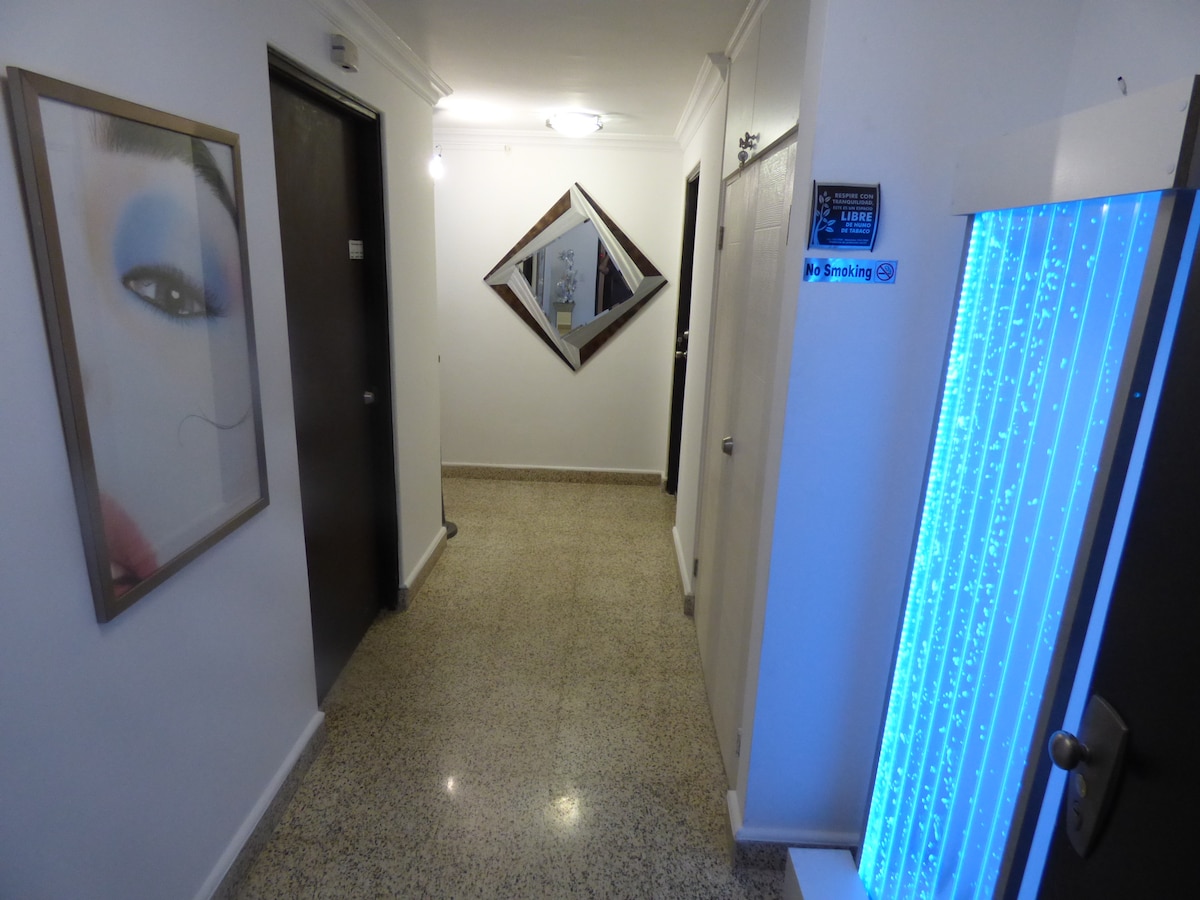 A well-lit corridor features glossy tile flooring and smooth walls, enhanced by a decorative mirror and framed artwork. The corridor is lined with multiple doors, leading to private spaces, while a blue accent light adds a calming touch.
