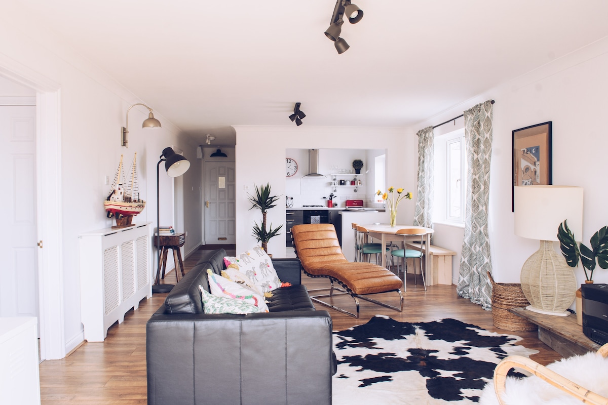 An open living area features a spacious seating arrangement with a dark leather sofa and a stylish armchair. A dining table is positioned near a window, surrounded by light. The kitchen is visible in the background, showcasing modern appliances and decorative elements including plants.
