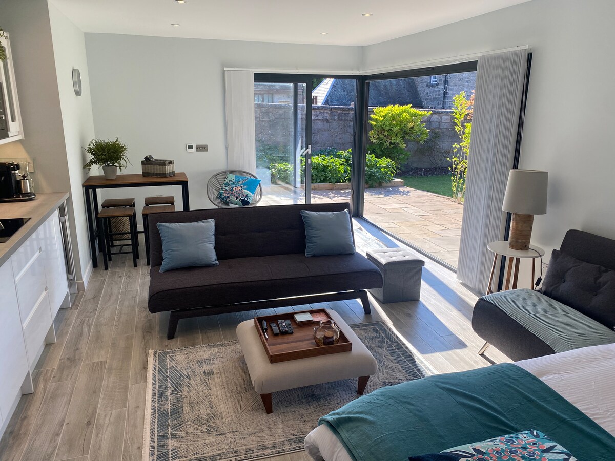 The living area includes a stylish sofa with soft pillows, a coffee table, and a dining bar with high stools. Bi-fold doors open to a private patio and garden. Natural light enhances the tranquil atmosphere, complemented by modern furnishings and decor.
