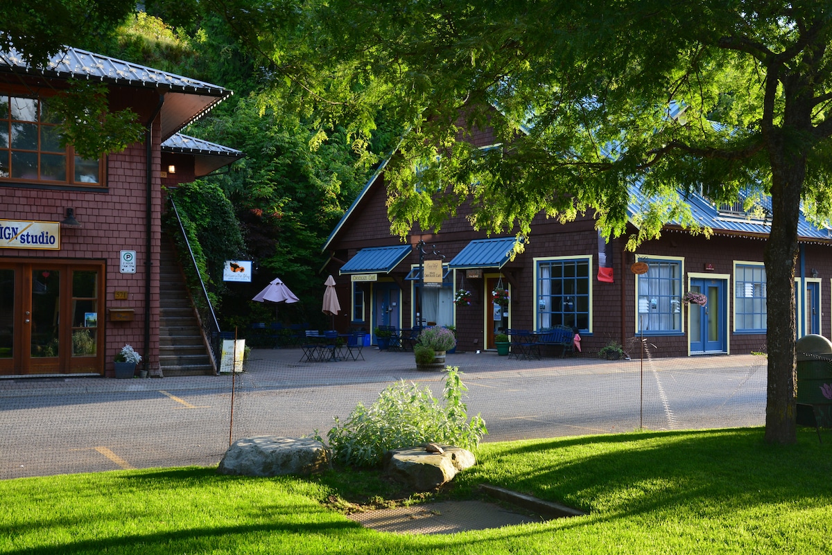A view of Artisan Square features rustic buildings with wooden facades under a clear sky. The area is enhanced by vibrant greenery and a well-maintained lawn, with seating options including blue chairs and umbrellas visible. Pathways and small stones create a welcoming atmosphere.