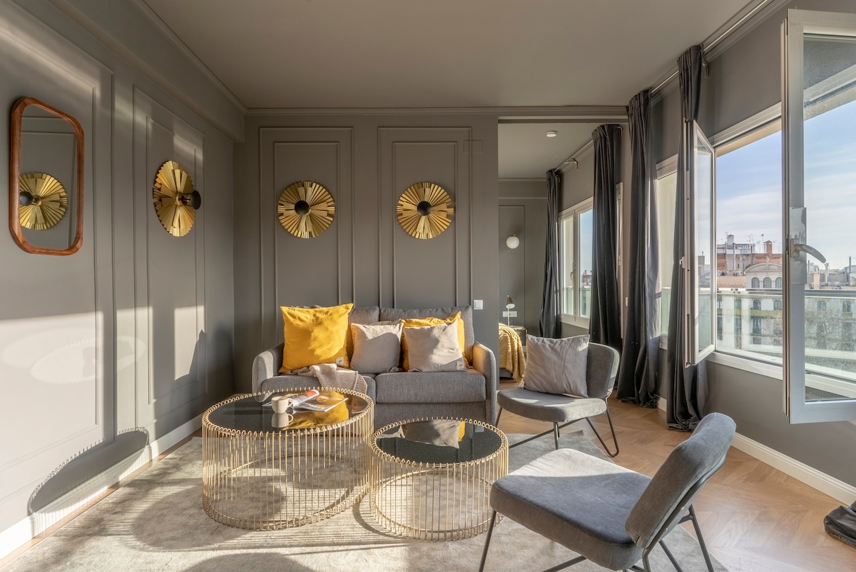 A cozy living area features a sofa adorned with decorative yellow cushions. Two elegant gold-accented tables sit atop a soft rug, while large windows bring in natural light. Stylish wall décor complements the ambiance, enhancing the modern design of the space.