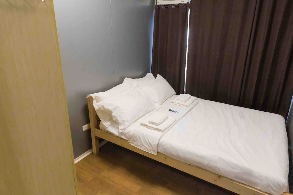 A compact room features a full double-sized bed with crisp, white linens. The walls are finished in a neutral tone, complemented by dark curtains that enhance privacy. Towels are neatly arranged on the bed, and a closet is visible in the corner.