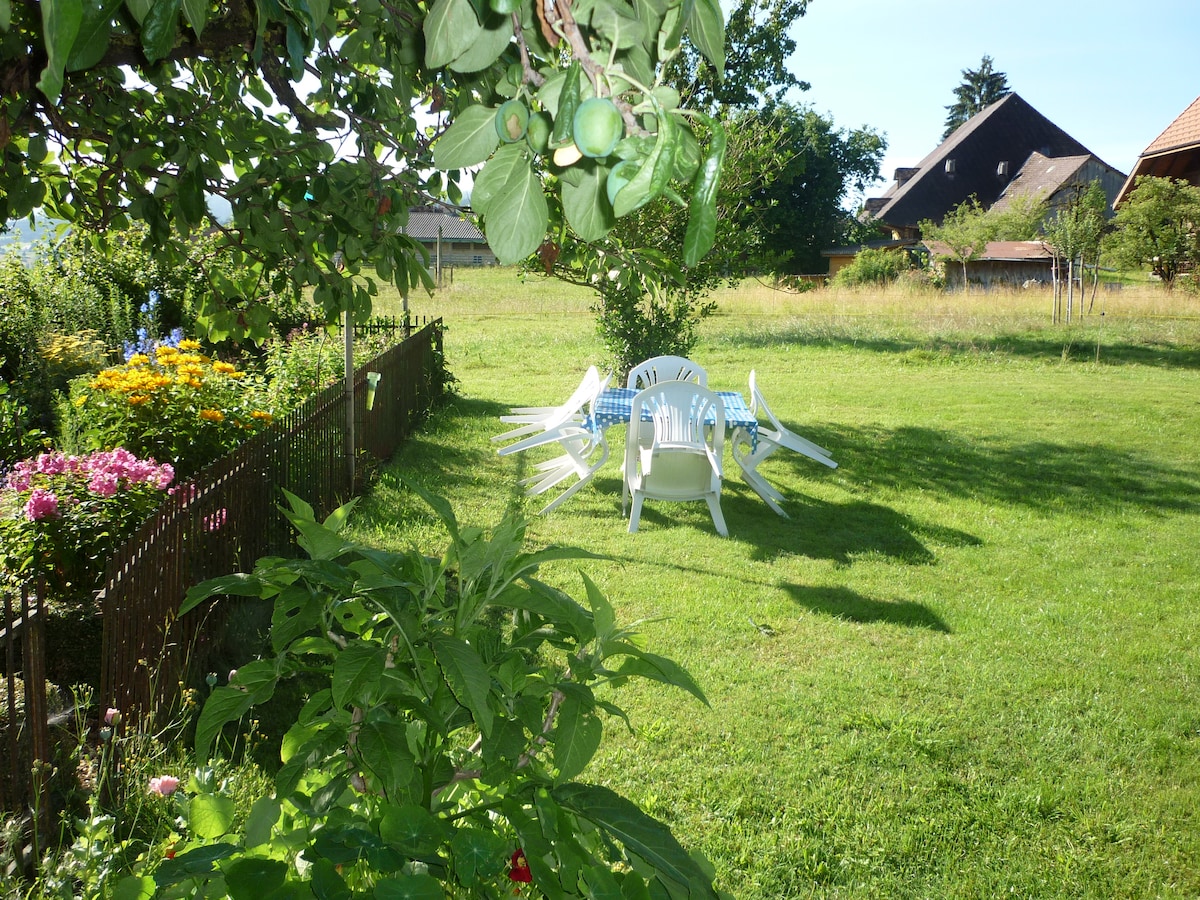 An expansive garden area is presented, featuring a round table surrounded by white plastic chairs. The lush green grass is bordered by colorful flowers and a wooden fence, with distant views of nearby structures partially obscured by trees.