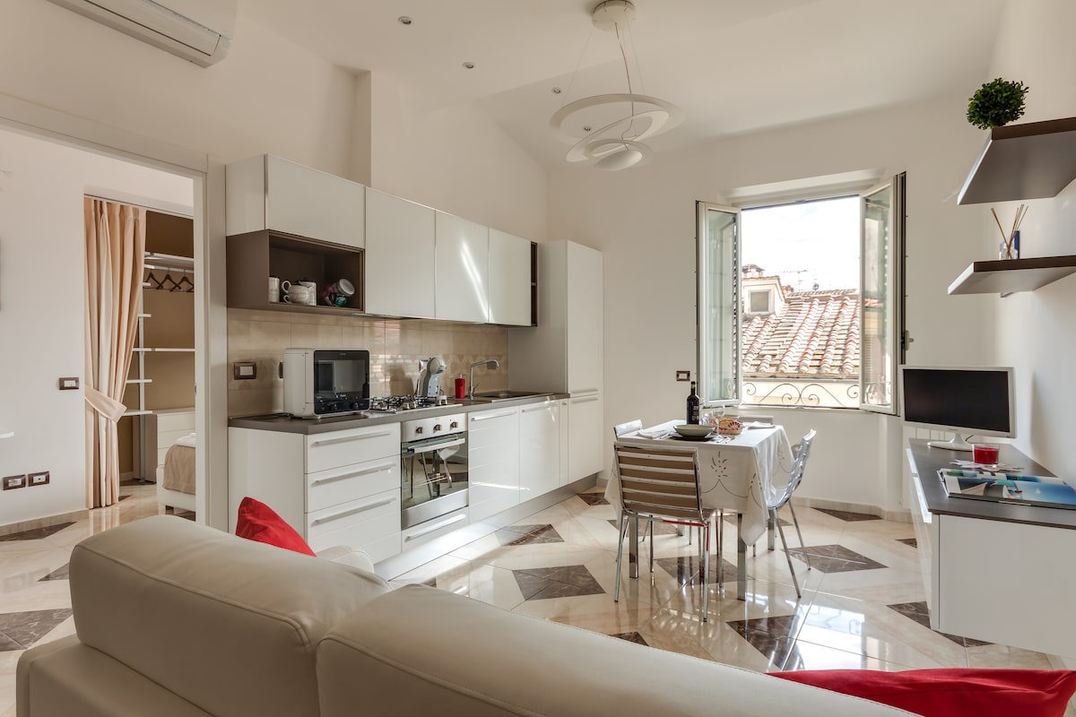 The modern kitchen area is equipped with stainless steel appliances and ample cabinet space. A white dining table with striped chairs is positioned near a window, offering views of the rooftop. The open layout seamlessly connects to the living area featuring a light-colored sofa.