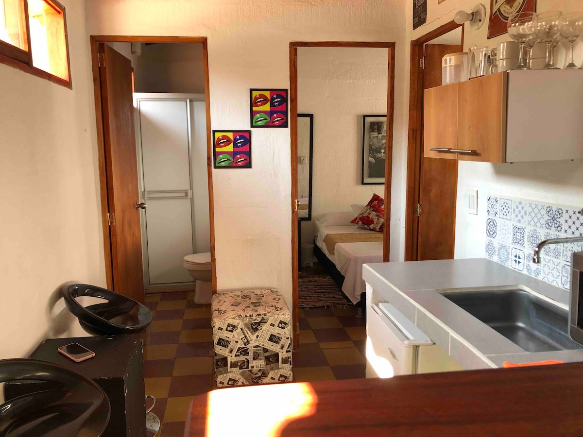 A compact kitchen area is equipped with a sink and countertop, adjacent to a dining space featuring a small patterned stool. Two wooden doors lead to separate rooms, while a bathroom is visible at the end of a short hallway. Colorful artwork adorns the wall.