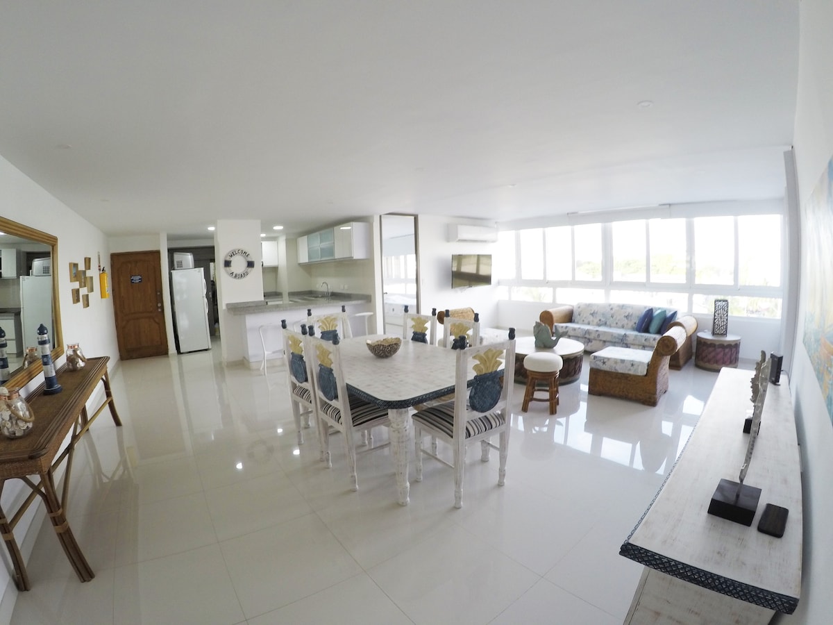 A spacious living and dining area features multiple large windows allowing natural light to fill the room. A comfortable sofa and armchair are arranged thoughtfully around a light-colored table, with elegant decor placed on the surfaces. A modern kitchen is visible in the background.