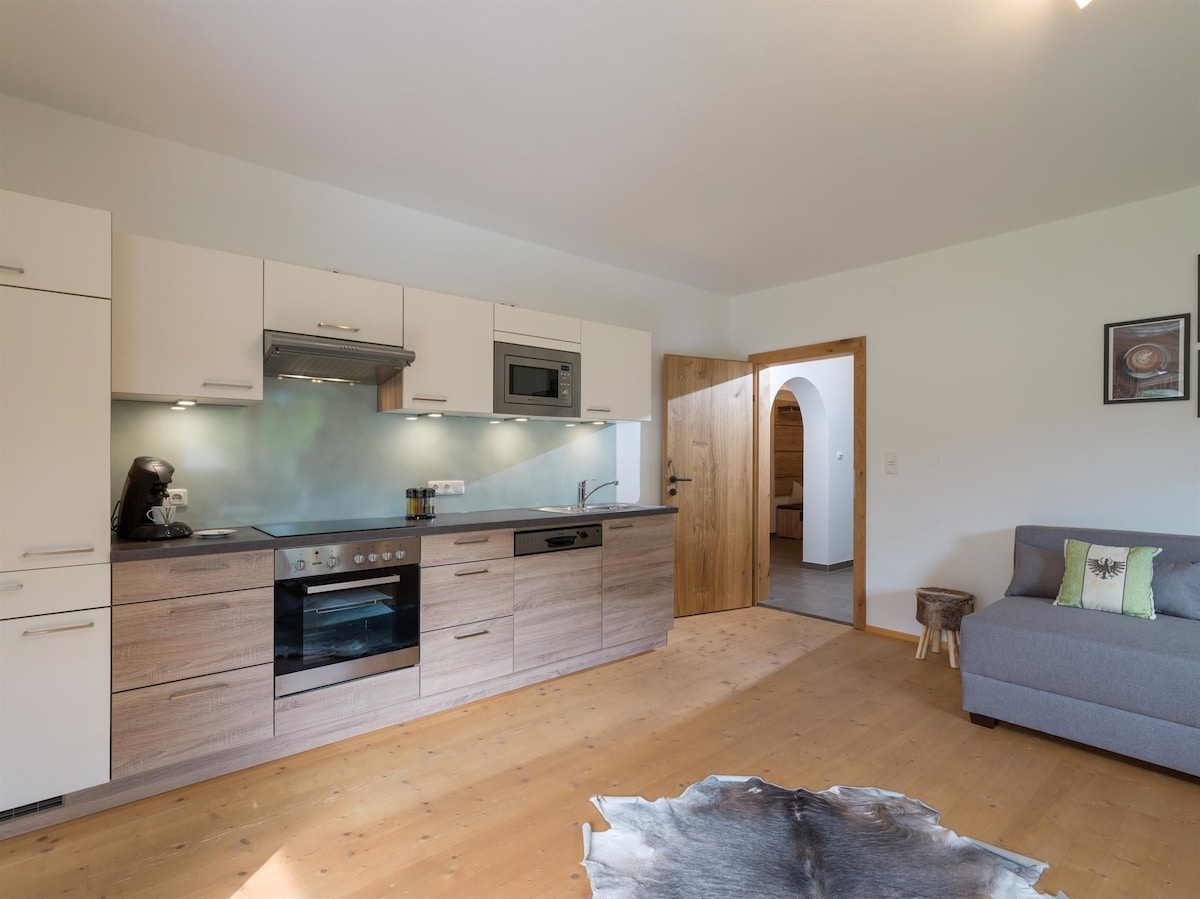 A modern kitchen area features sleek cabinetry and a stove with an oven beneath. A gray couch provides seating in the nearby living space, while a door leads to an adjacent room. Natural light enhances the warm wood flooring throughout the area.