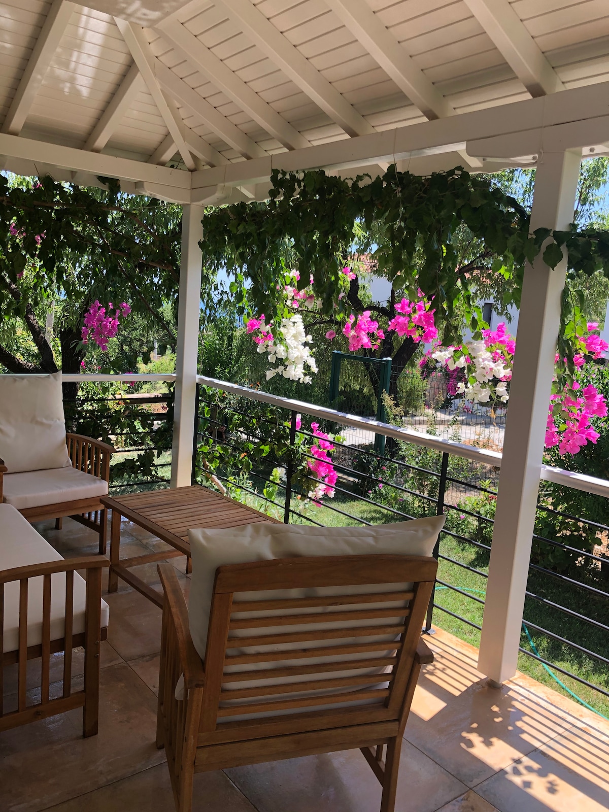A covered outdoor seating area is presented, featuring a wooden frame with light-colored cushions. Lush greenery and vibrant pink and white flowers hang from the trellis above, creating a serene atmosphere. Tiled flooring complements the natural surroundings.