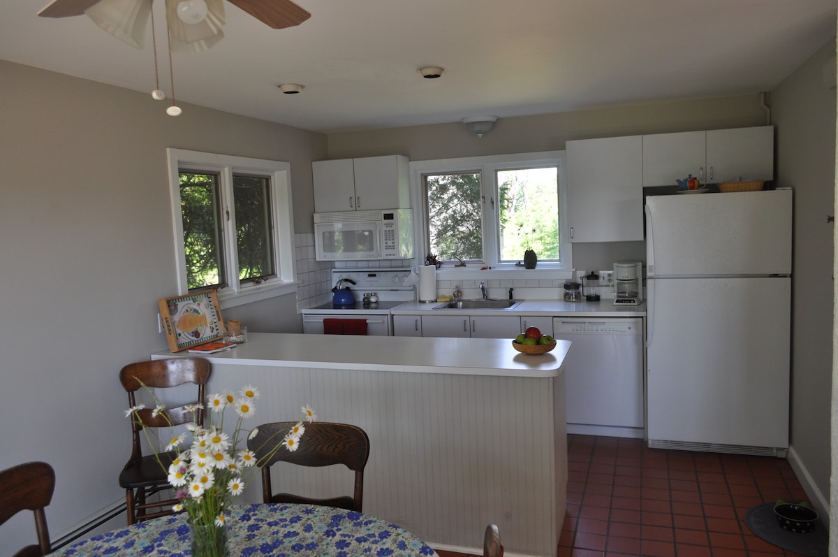 The kitchen features modern appliances, including a refrigerator and microwave, with ample counter space. Natural light filters through multiple windows, illuminating the area. A round table with floral tablecloth and chairs is positioned nearby, offering a cozy dining spot.