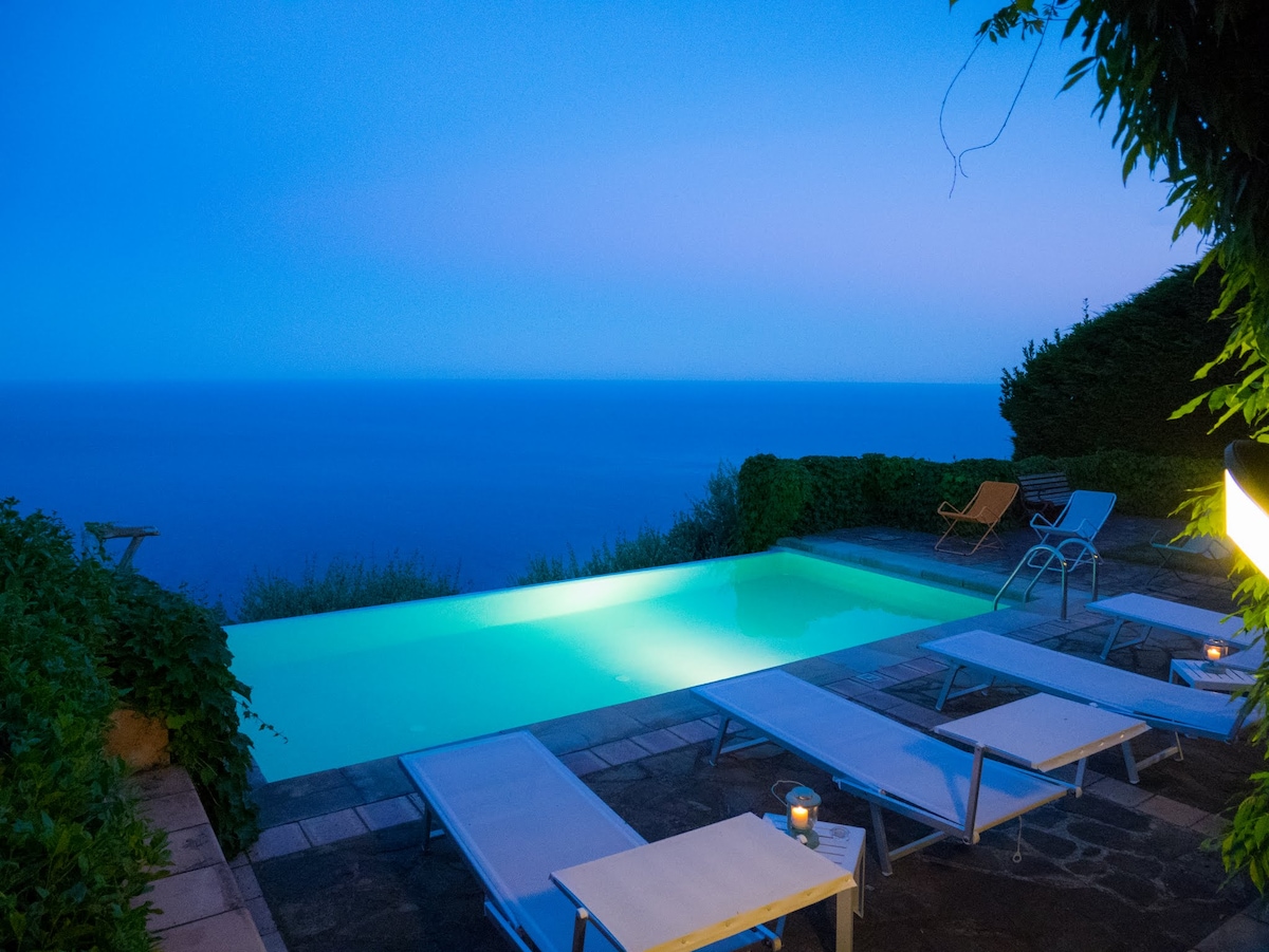 An infinity swimming pool is illuminated in soft turquoise under evening light, offering views of the sea. Surrounding the pool, loungers are arranged on a stone patio, complemented by green foliage and distant seating. The scene conveys a serene and tranquil atmosphere.