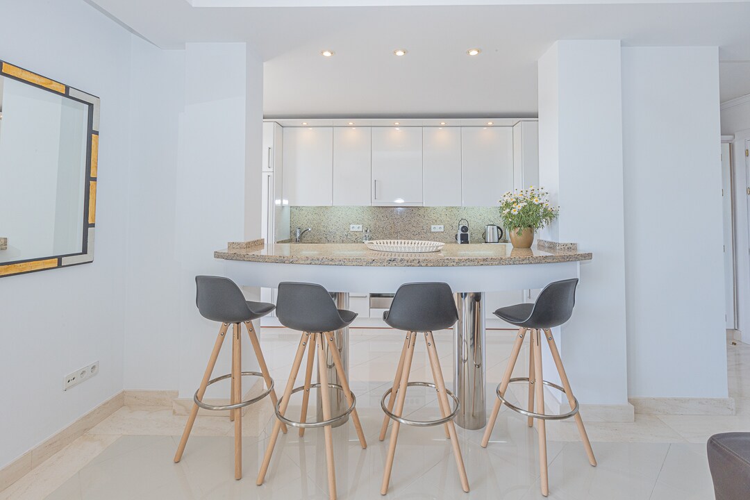 A modern kitchen area features a curved bar with four high stools, set against a backdrop of white cabinetry and light countertops. Soft lighting illuminates the space, highlighting the sleek design and clean lines, creating an inviting atmosphere for casual dining.