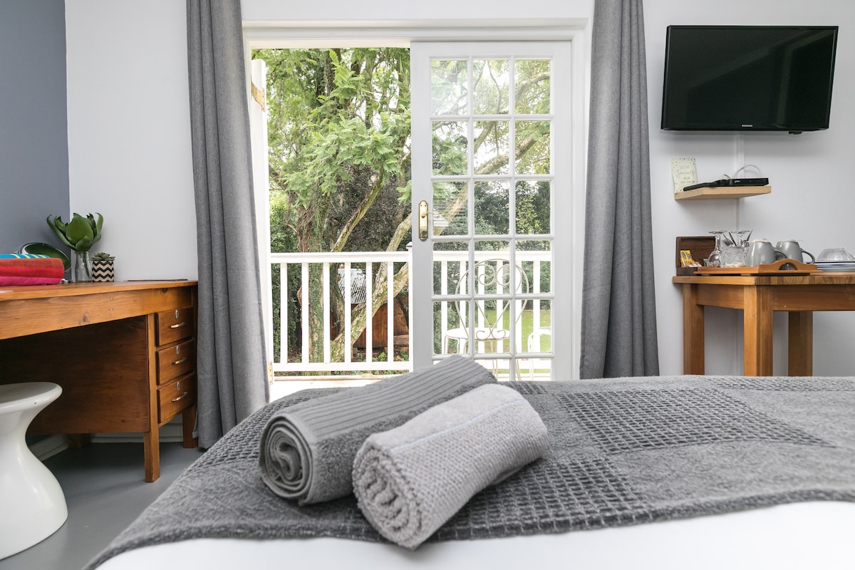 The image shows a light-filled bedroom featuring a spacious bed with neatly arranged grey towels. A flat-screen TV is mounted on the wall. Large glass doors open to a tranquil garden view, framed by soft grey curtains, adding to the room's modern aesthetic.
